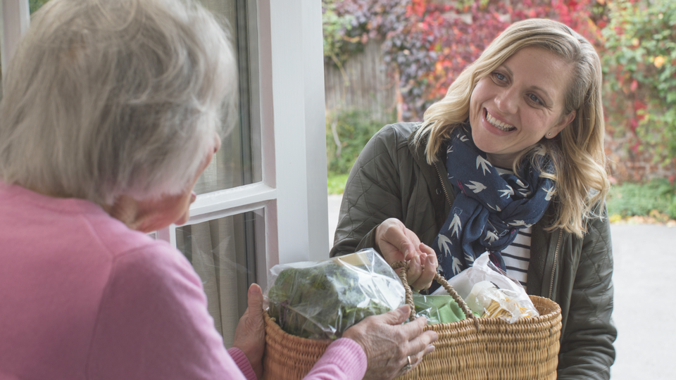 Smiling woman delivering a wicker basket of groceries to an elderly person at a doorway.
